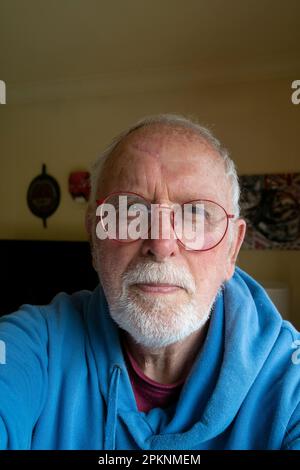 Ein 80-jähriger Mann, ein Rentner mit grauem Bart und roter Brille und blauem Kapuzenpullover Stockfoto