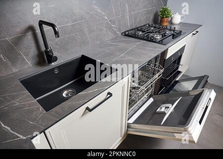 Zeitgenössische Küche mit eingebautem, geöffnetem Geschirrspülofen, quadratischem, schwarzem Gaskochfeld mit grauen Porzellan-Steingut-Ablageflächen und Schränken. Stockfoto