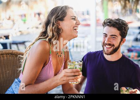 Eine Nahaufnahme einer freudigen, leicht brünetten Frau, die einen Mojito hält, mit einem bärtigen Mann an einer Strandbar sitzt, beide lachen heftig zusammen. Stockfoto