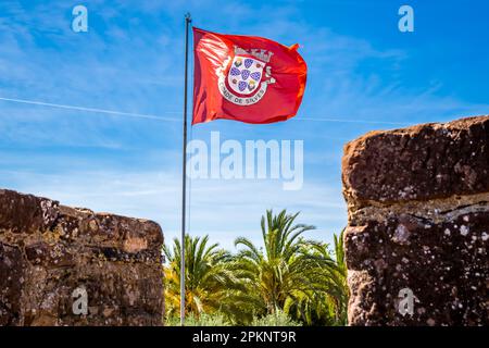 Niedriger Winkel Blick auf die rote Flagge von Cidade de Silves, geschmückt mit dem Wappen, flattern im Wind gegen den blauen Himmel, mit Zinnen. Stockfoto