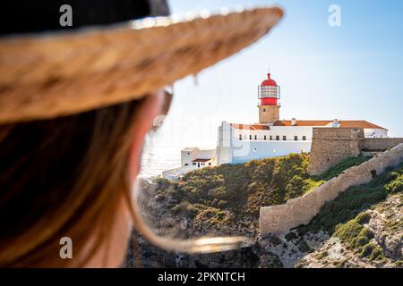 Rückansicht einer Touristenfrau mit Hut, die den atemberaubenden Blick auf den Leuchtturm Farol do Cabo de São Vicente hoch oben auf den zerklüfteten Klippen genießt. Stockfoto