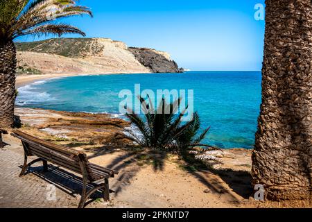 An einem sonnigen Maitag befindet sich eine leere Bank zwischen Palmen an der Luz Promenade und bietet einen ruhigen und ruhigen Blick auf den Strand Praia da Luz. Stockfoto