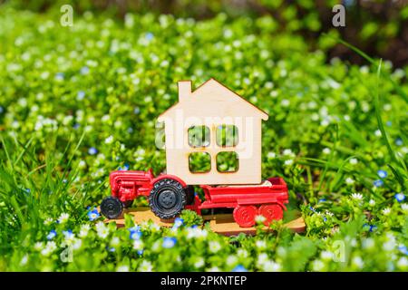 Die Essenz des Landlebens im Frühling, mit einem Spielzeugtraktor, der ein Miniatur-Flachhaus auf einem Blumenbeet aus grünem Gras und Colo trägt Stockfoto