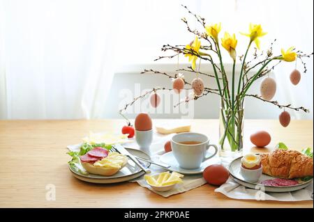 Das Osterfrühstück wird auf einem Holztisch mit einem Strauß aus Narzissen, Frühlingszweigen und hängenden Eierdekorationen, Kopierbereich, ausgewähltem Fokus serviert Stockfoto