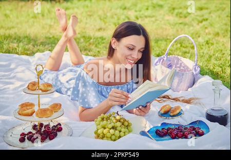 Picknickszene: Hübsches lächelndes Mädchen, das im Park auf einem Karo liegt und ein Buch liest. Außenporträt einer schönen, glücklichen jungen Frau in blauem Kleid Stockfoto