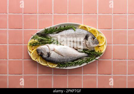 Dorada-Fisch mit Zitronenscheiben und Rosmarin-Zweigen in einem ovalen Gericht auf pinkfarbenem Fliesenhintergrund, Draufsicht, Kopierbereich Stockfoto