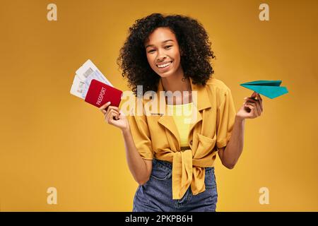 Internationaler Reisepass mit Flugtickets und Papierflugzeug in den Händen einer afroamerikanischen Frau, auf gelbem Hintergrund. Flugreisenkonzept Stockfoto