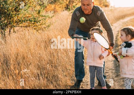 Dad bringt Triplet-Mädchen Tennis im Freien bei. Der Ball fliegt Stockfoto