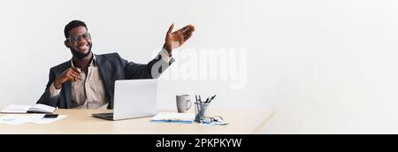 Glücklicher erfolgreicher afroamerikanischer Geschäftsmann, der am Tisch sitzt, mit einem Laptop, der die Hand auf einem leeren Raum zeigt Stockfoto