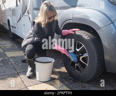 Eine attraktive Frau wäscht das Rad ihres Wohnwagens mit einer blauen Bürste. Sie trägt pinkfarbene Gummihandschuhe, schwarze Stiefel und Sonnenbrille.A Stockfoto