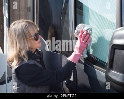 Eine attraktive Dame wischt mit einem Tuch das Seitenfenster ihres Wohnwagens ab. Sie trägt pinkfarbene Gummihandschuhe und eine Sonnenbrille Stockfoto