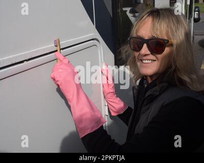 Eine attraktive lächelnde Frauenhand reinigt ihr Wohnmobil mit einer Zahnbürste. Sie trägt pinkfarbene Gummihandschuhe und eine Sonnenbrille. Zufrieden Stockfoto