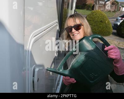 Eine attraktive lächelnde Dame, die in die Kamera schaut, schüttet Wasser auf ihr Wohnmobil mit einer grünen Gießkanne. Sie trägt einen rosa Gummihandschuh Stockfoto