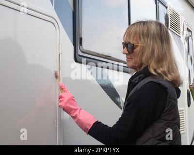 Eine attraktive lächelnde Frauenhand reinigt ihr Wohnmobil mit einem Schwamm. Sie trägt pinkfarbene Gummihandschuhe und eine Sonnenbrille. Zufrieden Stockfoto