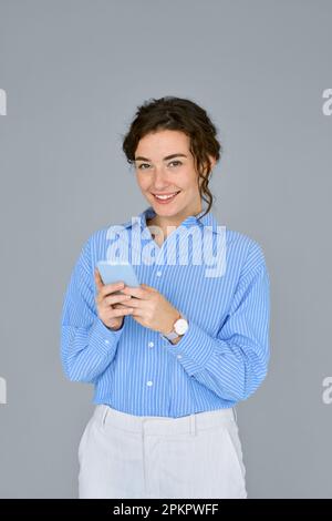 Junge, glückliche Geschäftsfrau, die ein Handy hält und an einer grauen Wand steht. Stockfoto