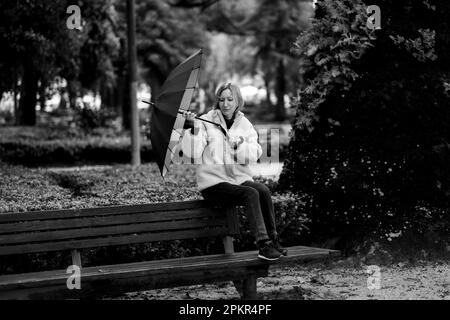 Eine Frau sitzt auf einer Parkbank, ein Regenschirm ist bei Regen kaputt gegangen. Schwarzweißfoto. Stockfoto