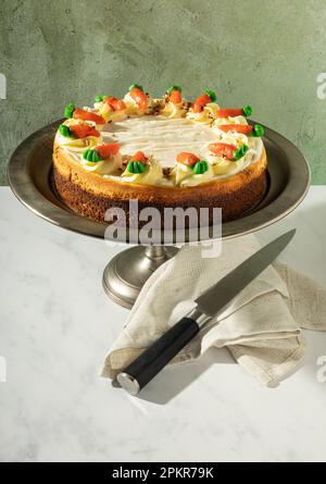 Käsekuchen mit voller Karotte auf einem Zinnkuchenständer, Messer und Stoffserviette im Vordergrund, auf grünem Hintergrund. Stockfoto