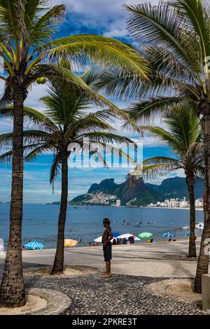 Afrikanischer brasilianischer Junge unter Palmen am Arpoador Strand - Favela do Vidigal ( Vidigal Slum ), Two Brothers Berg ( Morro Dois irmaos ) und Ipanema Strand im Hintergrund. Rio de Janeiro, Brasilien. Stockfoto