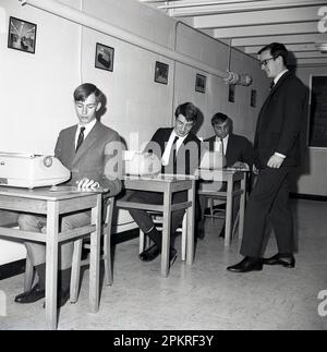 1960er, historische, gut gekleidete männliche Studenten, die an Schreibtischen mit elektrischen Schreibmaschinen sitzen und von einem jungen erwachsenen männlichen Lehrer oder Supervisor in einem Schreibkurs, Middletown, USA, beaufsichtigt werden. Modell der verwendeten Schreibmaschinen ist die IBM Selectric Schreibmaschine, die 1961 erstmals auf den Markt kam. Stockfoto