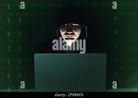 Hacker in der Kapuze sitzt nachts vor seinem Laptop, grüne Zahlen im Hintergrund. Cyberkriminelle Stockfoto