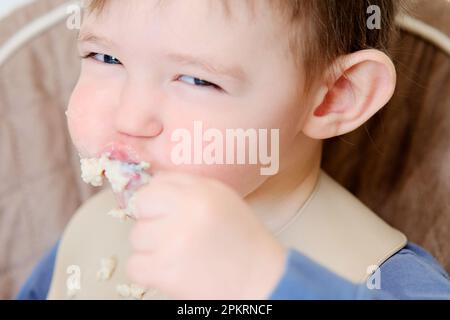 Ein glückliches Kind isst Haferbrei mit einem Löffel, während es auf einem Hochstuhl sitzt. Das Baby im Lätzchen isst Haferbrei selbst. Kind im Alter von etwa zwei Jahren (1 Jahre Stockfoto