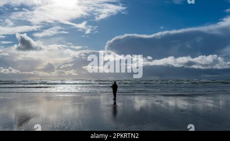 Ein alleinstehender Fischer, der bei Ebbe am Meer steht, sich im nassen Sandstrand der Küste spiegelt und von dem Sonnenlicht beleuchtet wird Stockfoto