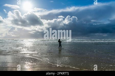 Ein alleinstehender Fischer in Neoprenanzug steht im Wasser der Ebbe und wird vom Sonnenlicht beleuchtet Stockfoto