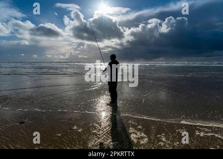 Ein alleinstehender Fischer in Neoprenanzug steht bei Ebbe am Meer, reflektiert im nassen Ufersand und beleuchtet vom Sonnenuntergang Stockfoto