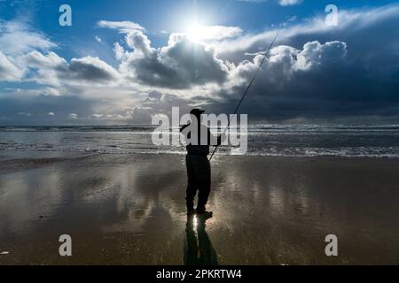 Ein alleinstehender Fischer in Neoprenanzug steht bei Ebbe am Meer, reflektiert im nassen Ufersand und beleuchtet vom Sonnenuntergang Stockfoto