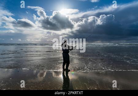 Ein alleinstehender Fischer in Neoprenanzug steht bei Ebbe am Meer, reflektiert im nassen Ufersand und beleuchtet vom Sonnenuntergang Stockfoto