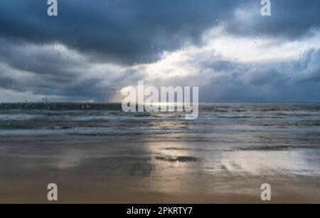 Bei stürmischem, windigem Wetter mit Sonnenuntergangsstrahlen, die durch die Wolken im Hintergrund fließen, werden die Regentropfen am Fenster zerquetscht Stockfoto