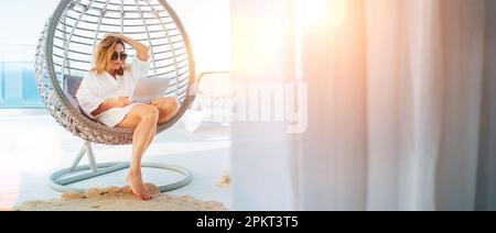 Eine wunderschöne Frau mittleren Alters, die in einem bequemen Hängestuhl auf einer sonnigen Terrasse mit einem modernen Laptop sitzt und im Internet shoppt Stockfoto