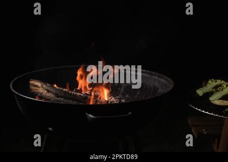 Brennholz brennt in einem runden Barbecue in der Nacht. Dunkler Hintergrund. Vorbereitung zum Kochen von Speisen auf dem Grill. Stockfoto