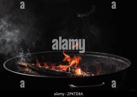 Brennholz brennt in einem runden Barbecue in der Nacht. Dunkler Hintergrund. Vorbereitung zum Kochen von Speisen auf dem Grill. Stockfoto