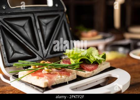 Sandwiches aus Schinken, Gemüse und Käse in einem Sandwichmaker machen. Zwei Toasts mit verschiedenen Füllungen – Nahaufnahme. Stockfoto