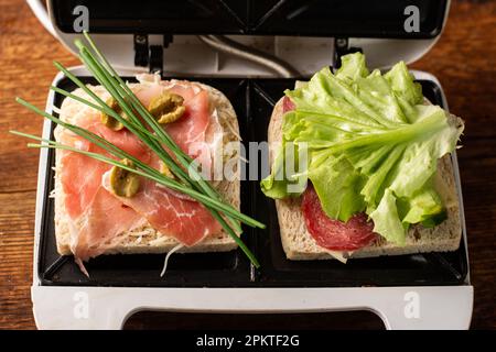 Sandwiches aus Schinken, Gemüse und Käse in einem Sandwichmaker machen. Zwei Toasts mit verschiedenen Füllungen – Nahaufnahme. Stockfoto