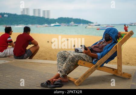 Pattaya, Thailand - Oktober 16,2022: Strand Ein junger Thailänder mit schwarzer Gesichtsmaske entspannte sich am Strand in einem Sitz. Stockfoto