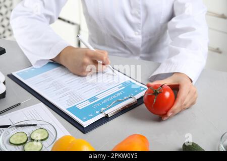 Wissenschaftler mit Labortestform und frischer Tomate am Tisch im Haus, Nahaufnahme. Gifterkennung Stockfoto