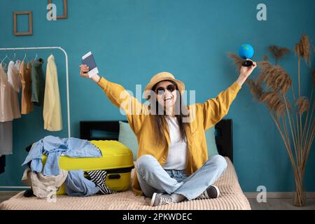 Junge, glückliche Reisende, die sich auf einen Sommerurlaub vorbereiten, mit ihrem hellgelben Koffer und ihrer Kleidung, die gepackt werden kann, zeigt Stockfoto