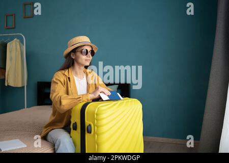 Junge hispanische Frau, die auf dem Bett saß und ihren Pass und Koffer hielt, bevor sie in den Urlaub ging, und durch das Fenster schaute. Speicherplatz kopieren Stockfoto