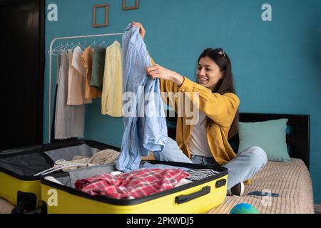 Fröhliche junge hispanische Frau packt Koffer, packt ihre Kleider, macht sich bereit für den Urlaub zu Hause. Millenial-Frau, die auf Touristenstour geht. Reisen Stockfoto