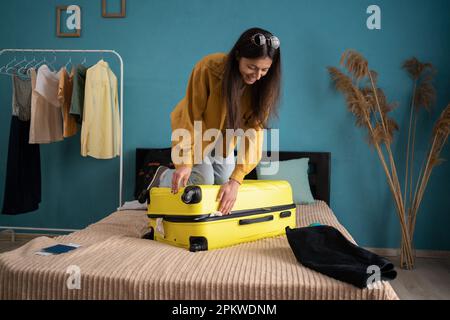 Vorbereitung des Reisekoffers zu Hause auf dem Bett. Eine junge Lateinin, die versucht, ihre Kleider in den gelben Koffer zu packen, um sich auf die Reise im Sommer vorzubereiten. Stockfoto