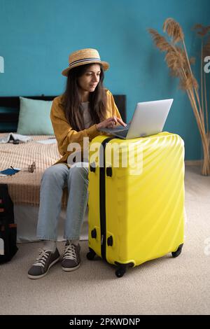 Eine Reisende Frau, die im Schlafzimmer lächelnd auf dem Bett sitzt, mit Trolleygepäck, während sie ihren Urlaub mit einem Laptop bucht, Sommerausflug online. Speicherplatz kopieren Stockfoto