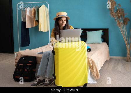 Eine aufgeregte Touristenfrau, die zu Hause einen Hut trägt, sitzt neben dem Koffer mit Laptop. Passagier, der am Wochenende ins Ausland reist. Flugflugkonzept. Stockfoto