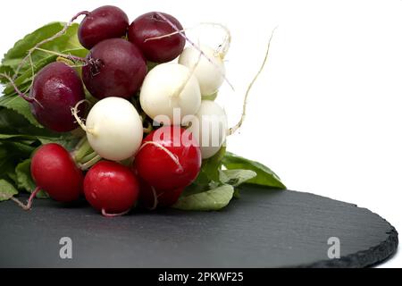 Frischer Rettich mit Blättern auf schwarzem Schieferteller auf weißem Hintergrund. Rettich (Raphanus sativus) Stockfoto