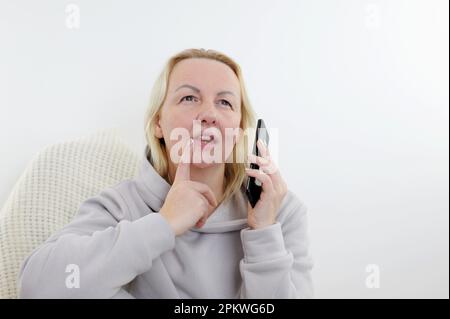 Träumige Frau mittleren Alters, die sich auf der Couch ruht, das Telefon in der Hand hält, auf einen Anruf wartet oder von Online-Chats in sozialen Netzwerken abgelenkt ist, ältere Frau lächelt, in der Ferne schaut, Fotos in der Freizeit genießt Stockfoto