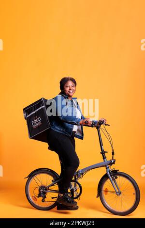 Pizzeria-Arbeiter, der einen Thermal-Rucksack mit sich führt, Fahrrad im Studio mit gelbem Hintergrund. Restaurantkurier bringt Pizza zum Mittagessen. Gastronomietransport Stockfoto