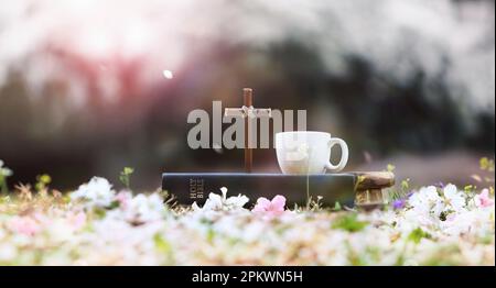 Frühlingshintergrund mit dem Kreuz Jesu Christi, der Bibel und einer Teetasse auf einem Blumenweg vor einem Kirschblütenwald an einem Frühlingstag Stockfoto