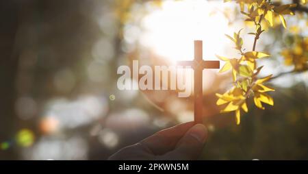 An einem warmen Frühlingstag mit starkem Sonnenlicht hält er gelbe Forsythien-Blumen und Knospen und das Heilige Kreuz Jesu Christi in seiner Hand. Stockfoto