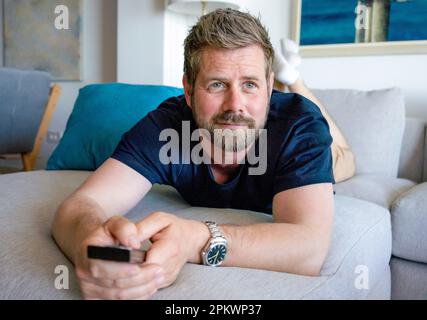 Freizeitaktivitäten am Wochenende. Weißer Mann der Generation Y mit Fernbedienung, der zu Hause Sender umschaltet. Ein glücklicher junger Mann, der interessante Progra sieht Stockfoto